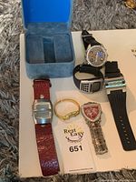 Assortment of 5 watches including digital, bracelet, shield face, studded large dial, and sporty black watch on carpet and white background.