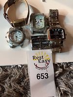 Five wristwatches displayed on a table, including metal and leather strap styles showing various dial designs and materials.