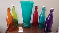 Full shot of the lot showing five coloured glass bottles arranged around a central turquoise glass vase on a wooden surface.