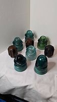 Nine glass and ceramic electrical insulators arranged on white fabric background. Colors include blue, green, and brown. Varied shapes and sizes.