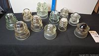 A group view showing 12 assorted glass insulators of various sizes and shapes on a black cloth surface, some with minor chips.