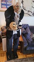 Full body view of Halloween butler figure holding skull tray standing indoors beside furniture.