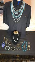 Photo of jewelry assortment on black shirt and black tablecloth, showing multi-strand turquoise and white bead necklace, leopard print pendant necklace, turquoise bead necklace, and multiple bracelets and earrings laid flat.