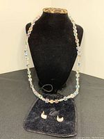 Full view of the Swarovski crystal bead necklace and crescent-shaped stud earrings displayed on a black stand with a black pouch beneath.