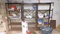 Three black metal 4-tier shelf units placed side by side in basement setting with concrete wall background. Various small household items on shelves, showing scratches and signs of wear.