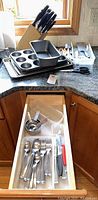 Photo shows baking pans including muffin tin, loaf pan, and cookie sheets stacked on countertop along with Henckels knife block and utensils. Drawer open with flatware and utensils organized in compartments.