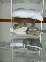 White metal shelving holding multiple folded bedding items including comforters, sheets, and pillowcases.