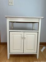 Front view of the white wood storage cabinet showing two cabinet doors with round knobs and an open shelf above.
