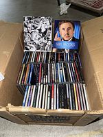 Full lot overview showing cardboard box containing Blu-ray sets and CDs arranged in rows inside.