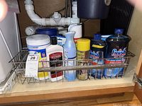 Under sink photo showing metal rack containing various cleaning supplies including Lysol disinfectant wipes, Weiman cooktop cleaner, Febreze air freshener, stainless steel cleaner, two bottles of Finish Jet Dry rinse aid, and a blue spray bottle.