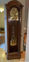 Full view of the tall brown wooden grandfather clock with glass front showing pendulum and weights.