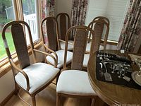 Six wooden dining chairs with woven wicker backs and white upholstered seats near window and table.