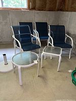 Four white metal-framed patio chairs with blue mesh seats arranged near two side tables with glass tops on a concrete floor.
