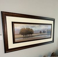 Framed landscape print featuring trees and water, surrounded by cream matting and a dark wood frame, hung on a wall.