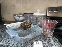 Photo showing a variety of bakeware and kitchen items including rectangular Pyrex baking dishes, a brown Anchor custard dish, glass and plastic measuring cups, salad spinner, metal strainers, and ladle on kitchen countertop.