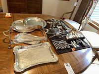 Full view of all items including silver plated serving trays, stainless steel flatware neatly arranged on a black cloth, and pewter cordial set on a wooden table.
