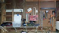 Photo showing a pegboard wall with hanging levels, clamps, a caulking gun, and a crowbar, with tools also placed on shelf: electric drill, socket wrenches, saws, and a step stool below.