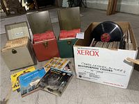 Photo shows several music record cases in beige, green, and red with 45 RPM vinyl singles in paper sleeves and a white Xerox box filled with loose vinyl records and record covers on concrete floor.