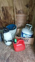 Wide shot showing chemical sprayer, propane tank, gasoline container, and two pails inside a wooden shed.