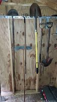 Photo showing six vintage garden tools hanging on a rack inside a shed: two shovels (pointed and rounded), a pitch fork, a garden rake, an edger, and a blue broom/brush.
