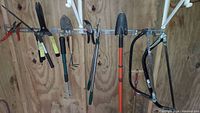 Photo showing various garden tools arranged on wall mounts including two hand bow saws, shovels, cultivator fork, pruners, and hedge trimmers.