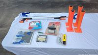 Wide shot of lot items arranged on white tablecloth outdoors showing picker uppers, wall mounts, filter, level tool, blade sharpener, and small packages