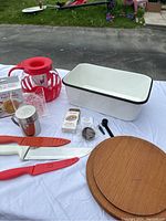 Photo showing red microwave popcorn popper, large white enamel pan, kitchen knives with red handles, stainless steel salt shaker, measuring spoon, reusable silicone lid, wooden plate, and grater on a white cloth.