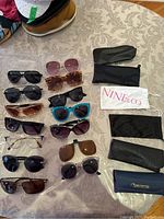 Top-down view of assorted sunglasses and eyeglasses arranged on tablecloth with several soft and hard cases on right.