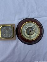 Two weather instruments on a white cloth background: the round wooden and brass barometer and the square Taylor Humidiguide hygrometer.