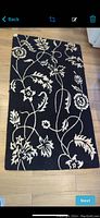 Full view of the rectangular black wool rug with a cream floral design placed on wooden floor.
