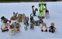 Miniature collectable figurines arranged on white surface including Raggedy Ann & Andy dolls, pewter animals, and blown glass animal figurines