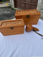 Photo of two wooden storage boxes and one carved wooden gourd vessel displayed on a white cloth outdoors, showing the front and sides with bronze clasps and woven top inserts.