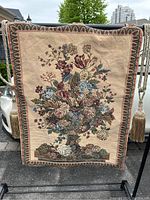 Full view of the tapestry wall hanging showing the floral pedestal planter design, hanging rod, and side tassels.