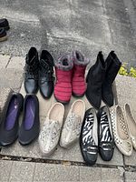 Seven pairs of ladies shoes and boots arranged in two rows on pavement, including boots and flats in various colors and materials.