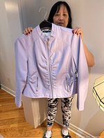 Front view of mauve fitted jacket on hanger held by woman, showing silver zipper pockets, ruching down center and sides, long sleeves, and short length.