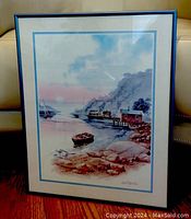 Framed watercolor painting showing a coastal scene with a boat, houses, and rocky shore under a cloudy sky.