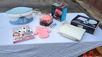 Photo shows entire lot items arranged on table outdoors with a white cloth. Items include a blue OBUSforme paraffin unit, pink soft hair rollers, boxed Conair soft rollers, white Conair bonnet dryer, cosmetic brush cleaning glove, and box for Conair bonnet dryer.