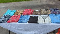 Lot layout of 13 women's shirts in various colors and patterns spread on a white surface outdoors