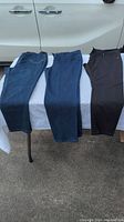 Three pairs of women's denim pants laid side by side on a white table: two blue pairs on left and middle, and one black pair on right. The photo shows full length of pants and waistband details.