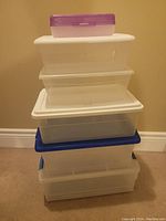 Stack of over 10 plastic storage containers in different sizes and colors; mostly clear with white and blue lids and one purple lid on top