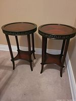 Two round wooden side tables with patterned inset tops, decorative carved edges, and lower shelves on carpeted floor.