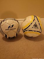 Photo showing two used soccer balls, one Pro Touch and one Candea, both uninflated and resting on carpeted floor against a beige wall.