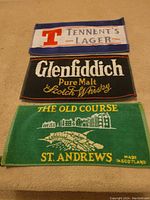 Three bar towels featuring Tennent's Lager, Glenfiddich Pure Malt Scotch Whisky, and The Old Course St. Andrews designs laid out flat on a surface.