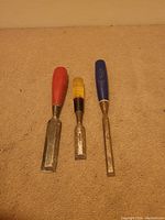 Three woodworking chisels placed on carpet background showing varying handle colors (red, yellow, blue) and blade widths with signs of rust and wear on metal blades.