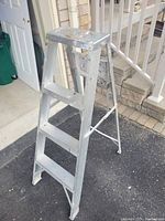 4 foot aluminum step ladder with four steps and a built-in shelf, placed outdoors on pavement near a porch and open door.