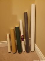 Photo showing six cylindrical shipping tubes in various colors and lengths against a beige wall and carpeted floor.