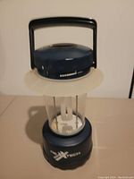 Front view of a blue X-Tech portable camp lantern with a black handle, battery level indicator, and clear light bulb housing.