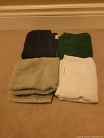 Four folded dish cloths in dark blue, forest green, beige, and white colors on the floor carpeted background.