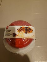 Top side view of red ceramic glazed brie baker with label band showing usage example of baked brie with toppings.