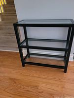 Black frame hall table with three clear glass shelves, placed on hardwood floor near stairs.